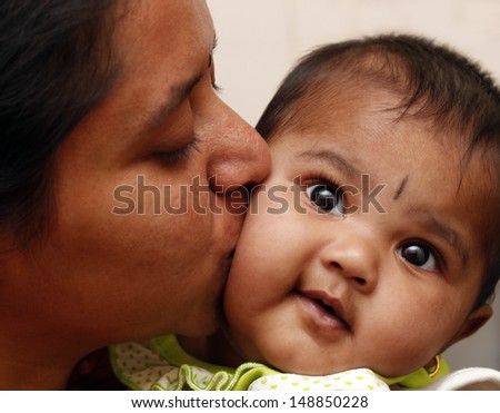 mother expressing love to daughter by kissing the child. The photo shows indian female kid smiling as her mummy embraces the toddler affectionately and kisses her