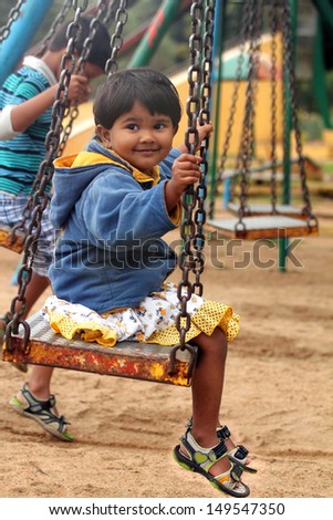 Beautiful &  happy indian girl ( child ) playing on a swing in a park. The photo shows summer time playground with a female kid smiling and sitting on a swing with a boy also playing in the background