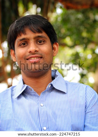 happy young indian man smiling and standing outdoors looking at the camera and very relaxed with blurred green tree leaves in the background