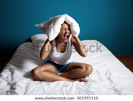 woman sitting on a bed and screamin, close her ears with a pillow