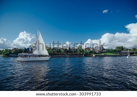 View from the board of a sailing yacht on the waters, sailing ships and the forest growing along the coast, as well as people\'s homes. Sailing yacht sails past the dock at private homes.