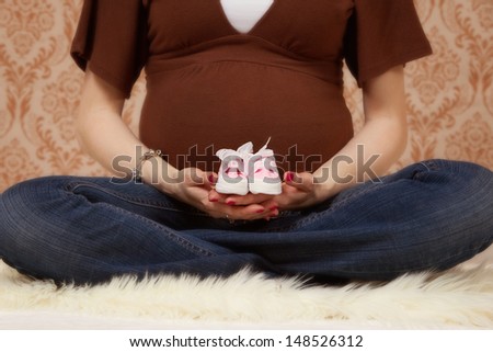 An expecting mother sitting crossed legged holding pink baby shoes with a soft defocused background