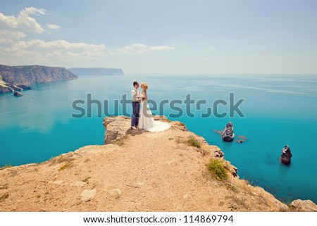 wedding theme: the bride and groom standing on a cliff near the azure waters. Groom in sunny summer day at the top of the bride hugging the mountains.