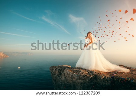 Bride in wedding dress stands on a cliff with a beautiful sea view from the top and flying confetti in the shape of hearts