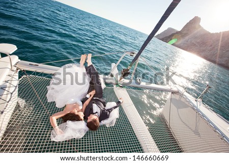 Wedding Couple Relaxing On Catamaran. Bride Lying On A White Wedding Dress, Groom In A Dark Suit