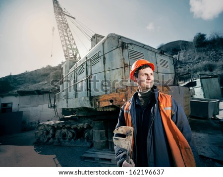 Worker In A Helmet With A Wrench In His Hand Standing Near A Construction Crane Lifting