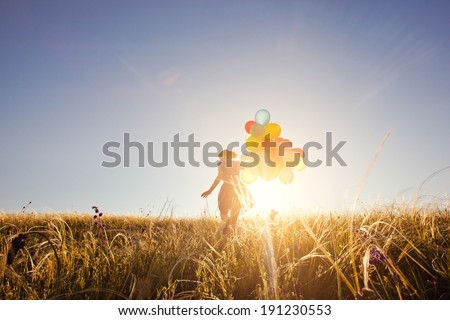 Happy woman with balloons running on the green field at sunset.