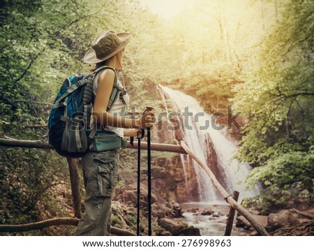 Hiking. Hikers woman with a backpack and a hat looking at a waterfall in the forest.