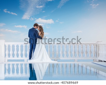 Wedding couple hugging and standing with her back outdoors near the pool. Happy bride and groom in love on their honeymoon. Woman in an elegant dress a man in the suit.