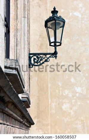 Old black wrought iron outdoor street lamp on mottled limestone wall in Europe. Modern electric bulb, cobwebs and dust show inside glass casing.