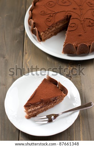 Piece of chocolate cake on a wooden table with the whole cake in the background