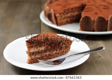 Piece of chocolate cake on a wooden table with the whole cake in the background