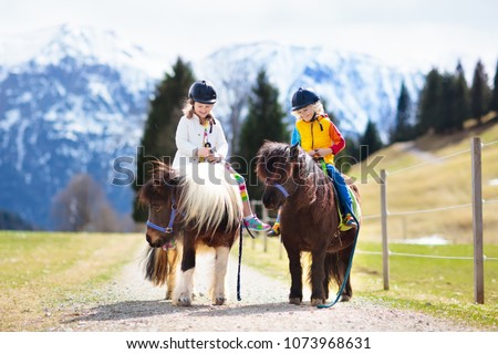 Kids riding pony in the Alps mountains. Family spring vacation on horse ranch in Austria, Tirol. Children ride horses. Kid taking care of animal. Child and pet. Little girl and boy in saddle on pony.