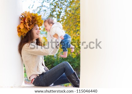 Attractive young woman with maple leaves wreath sitting between two white columns and playing with her baby