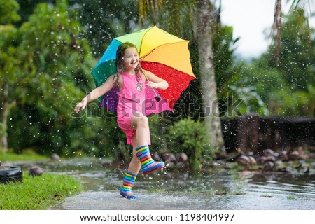 Kid playing out in the rain. Children with umbrella and rain boots play outdoors in heavy rain. Little girl jumping in muddy puddle. Kids fun by rainy autumn weather. Child running in tropical storm.