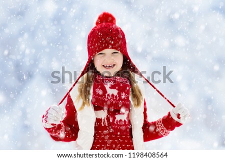 Child in red hat playing in snow on Christmas vacation. Winter outdoor fun. Kids play in snowy park on Xmas eve. Little girl in knitted sweater, scarf and mittens with Christmas decoration.