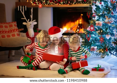 Children at Christmas tree and fireplace on Xmas eve. Family with kids celebrating Christmas at home. Boy and girl in matching pajamas decorating xmas tree and opening presents. Holiday gifts for kid.