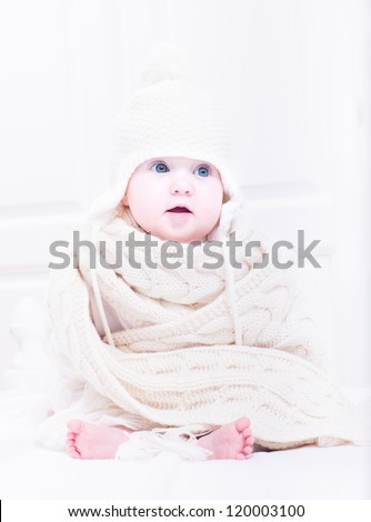 Sweet baby sitting in a white nursery in a huge hat and knitted scarf