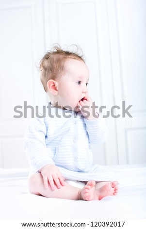 Sweet baby girl sucking on her finger sitting in a white nursery wearing a blue knitted dress