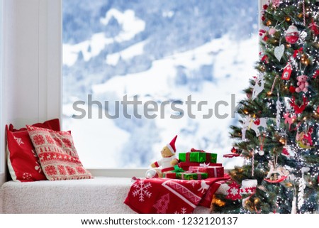 Christmas tree at window with view to snow and Swiss Alps mountains in winter. Decorated living room with Xmas gifts and presents for kids, pillows and toys. Family home seasonal interior decoration.