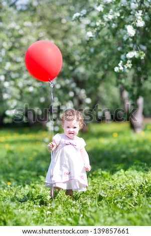 Adorable baby girl playing with a red balloon in a blooming garden
