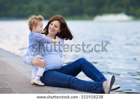 Young pregnant mother and her little baby daughter relaxing at a river bank in a city center