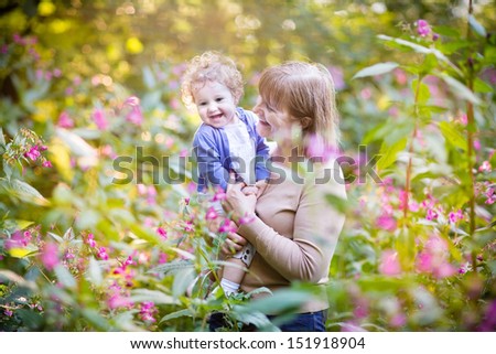Beautiful woman playing with a laughing baby girl on sunset in a garden with pink and red flowers