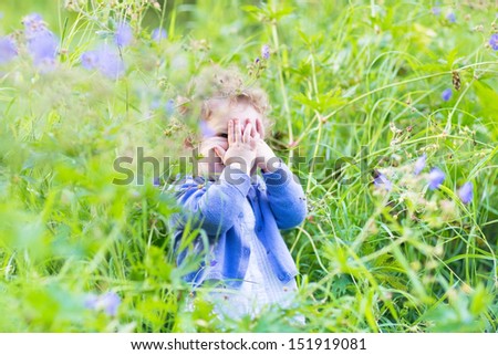 Cute funny baby girl playing hide and seek in the garden with blue and purple flowers