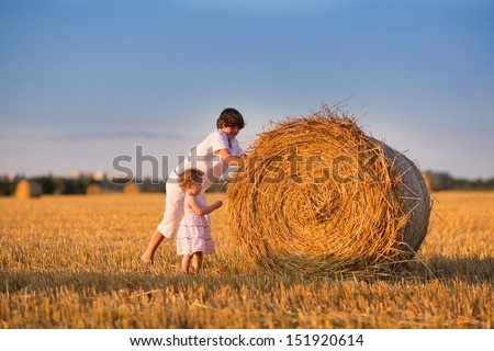 Brother and baby sister pushing hay bales in a field at sunset