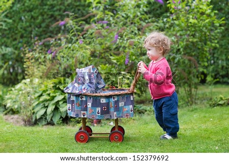 Funny curly baby girl playing with a vintage doll stroller in the garden