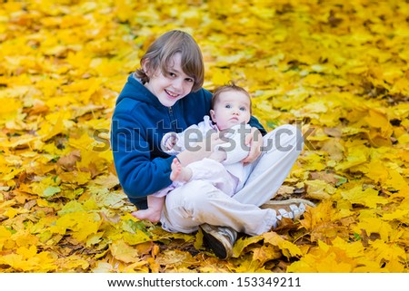 Cute brother holding his baby sister playing between yellow maple leaves