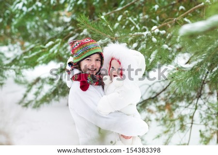 Cute boy hugging his baby sister in a beautiful winter park under a Christmas tree in snow both wearing warm white jackets and colorful knitted hats