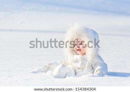 Adorable laughing baby girl in a warm white snow suit playing in snow on a very sunny and clear winter day in a park
