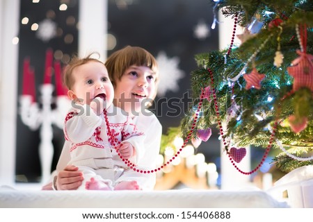 Beautiful portrait of a brother and baby sister in a dark living room next to a Christmas tree with candles and lights