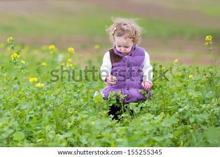 Cute little baby girl in a warm purple jacket playing with flowers in a field on a cold autumn day