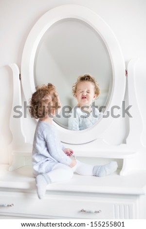 Very funny baby girl with curly hair looking at her reflection in a beautiful white bedroom with a classic dresser with a round mirror