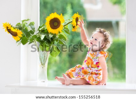 Happy smiling baby girl playing with beautiful sunflowers on a sunny summer day sitting at a window to the garden