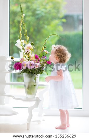 Adorable baby girl with curly hair wearing a white dress smelling beautiful flowers next to a wind and door to the garden