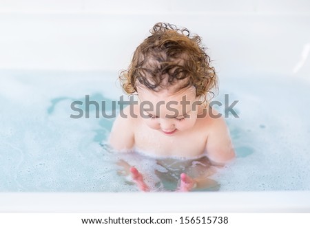 Adorable baby girl with curly hair playing with soap foam in a bath tub
