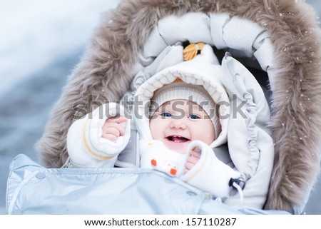 Happy Laughing Baby Girl Enjoying A Walk In A Snowy Winter Park Sitting In A Warm Stroller With Sheepskin Hood