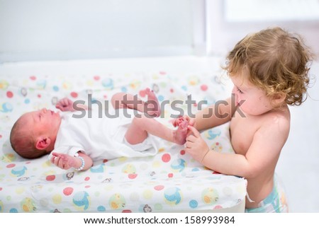 Portrait of a cute curly toddler girl playing with the feet of her newborn baby brother on a changing table