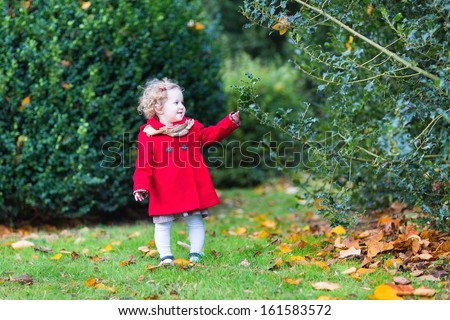 Adorable little toddler girl with curly hair wearing a red coat and a dress playing in a beautiful autumn park