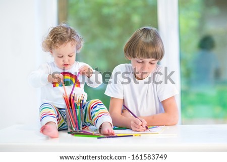 Funny toddler girl playing with colorful pencils sitting on a white desk next to a window watching her brother drawing