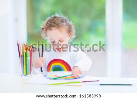 Sweet funny toddler girl painting a rainbow in a white room with a big window into the garden