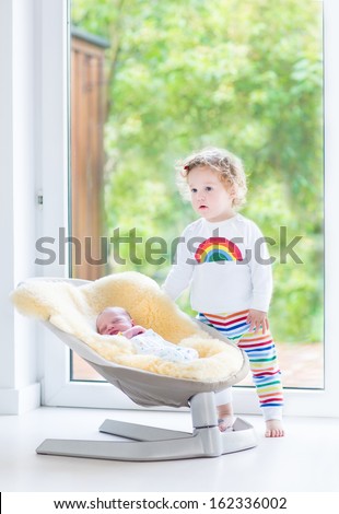 Cute toddler girl playing with her newborn baby brother relaxing in a swing next to a big window and door to the garden