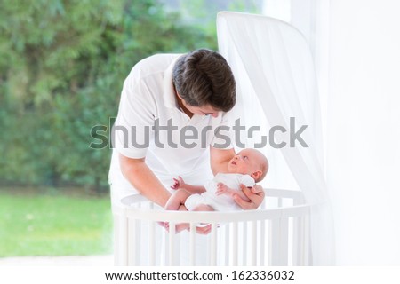 Young father putting his newborn baby into a white round crib with canopy next to a big window into the garden with green trees