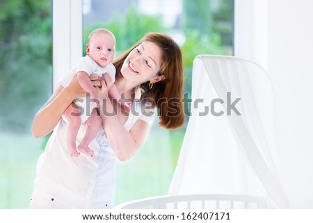 Young mother holding her newborn baby at a white round crib next to a big window