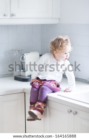 Adorable Toddler Girl Sitting In A White Kitchen On The Counter Top