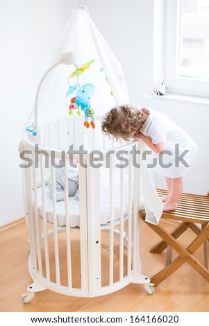 Funny curly toddler girl looking at her newborn baby brother sleeping in a white round crib with colorful toys and canopy