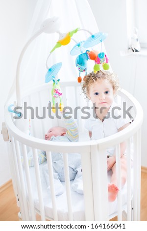 Cute Funny Toddler Girl Sitting In A Crib Of Her Her Newborn Baby Brother With Colorful Toys And Canopy